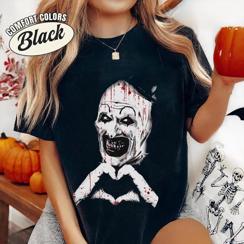 May include: Black t-shirt with a graphic of a scary clown face and a heart-shaped hand gesture. The text "Comfort Colors Black" is in a white oval on the upper left. The shirt is part of a Halloween-themed collection.