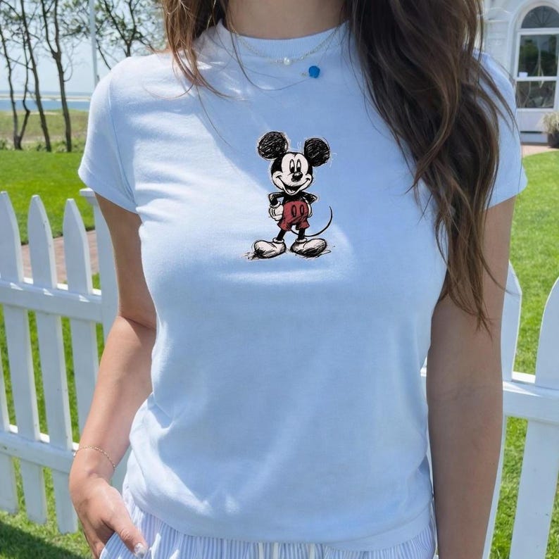 May include: Light blue t-shirt featuring a classic Mickey Mouse graphic. The cartoon character is depicted in black, red, and white. The shirt is a crew neck style and appears to be made of a soft material.