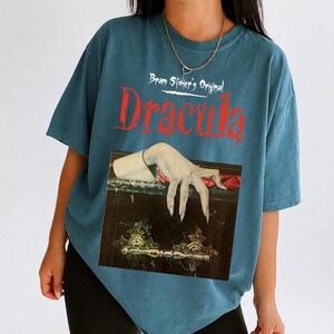 以下が含まれることがあります： 白と赤の文字で「Bram Stoker's Original Dracula」と書かれたスレートブルーの半袖Tシャツ。テキストの下には、暗く装飾された棺の縁に置かれた、長い爪を持つ青白い手のグラフィックが描かれています。