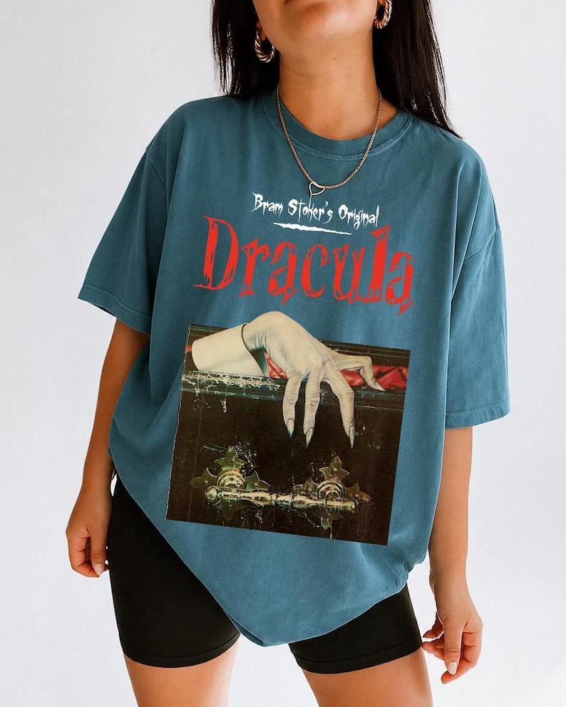以下が含まれることがあります： 白と赤の文字で「Bram Stoker's Original Dracula」と書かれたスレートブルーの半袖Tシャツ。テキストの下には、暗く装飾された棺の縁に置かれた、長い爪を持つ青白い手のグラフィックが描かれています。