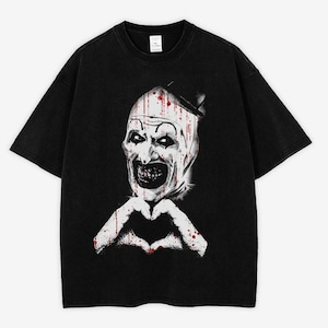 May include: Black t-shirt featuring a graphic of a scary clown with a white face, blood-like streaks, and a heart-shaped hand gesture. The clown has a sinister grin and is wearing a small hat. The shirt is made of a soft material.