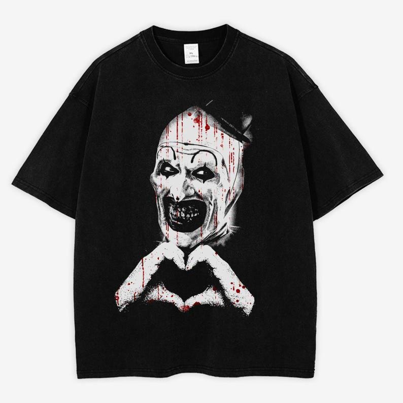 May include: Black t-shirt featuring a graphic of a scary clown with a white face, blood-like streaks, and a heart-shaped hand gesture. The clown has a sinister grin and is wearing a small hat. The shirt is made of a soft material.