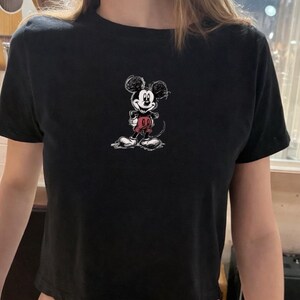 May include: Black crop top featuring a sketched Mickey Mouse graphic in black, red, and white. The cartoon character is centered on the front of the shirt. The top has short sleeves and a crew neck.