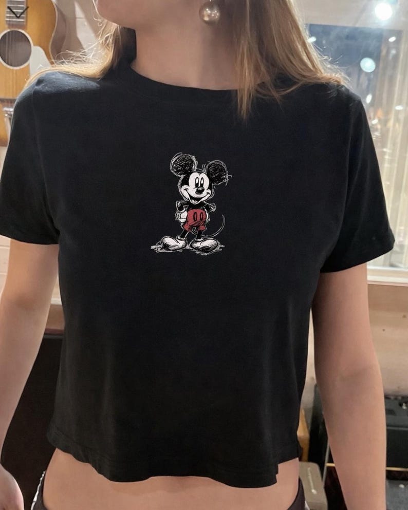 May include: Black crop top featuring a sketched Mickey Mouse graphic in black, red, and white. The cartoon character is centered on the front of the shirt. The top has short sleeves and a crew neck.