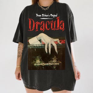 以下が含まれることがあります： 赤と白の文字で「Bram Stoker's Original Dracula」と書かれた黒の半袖Tシャツ。グラフィックには、装飾的な金属製の金具が付いた暗い棺桶から、長い爪を持つ青白い手が伸びている様子が描かれています。