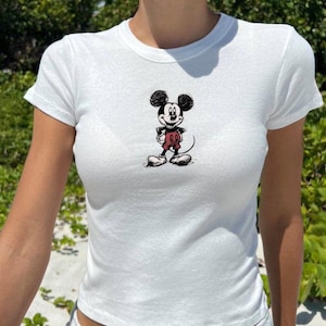 May include: White, short-sleeved t-shirt featuring a sketch-style Mickey Mouse graphic on the front. The cartoon character is depicted in black, red, and white. The shirt is made of a soft, lightweight material.