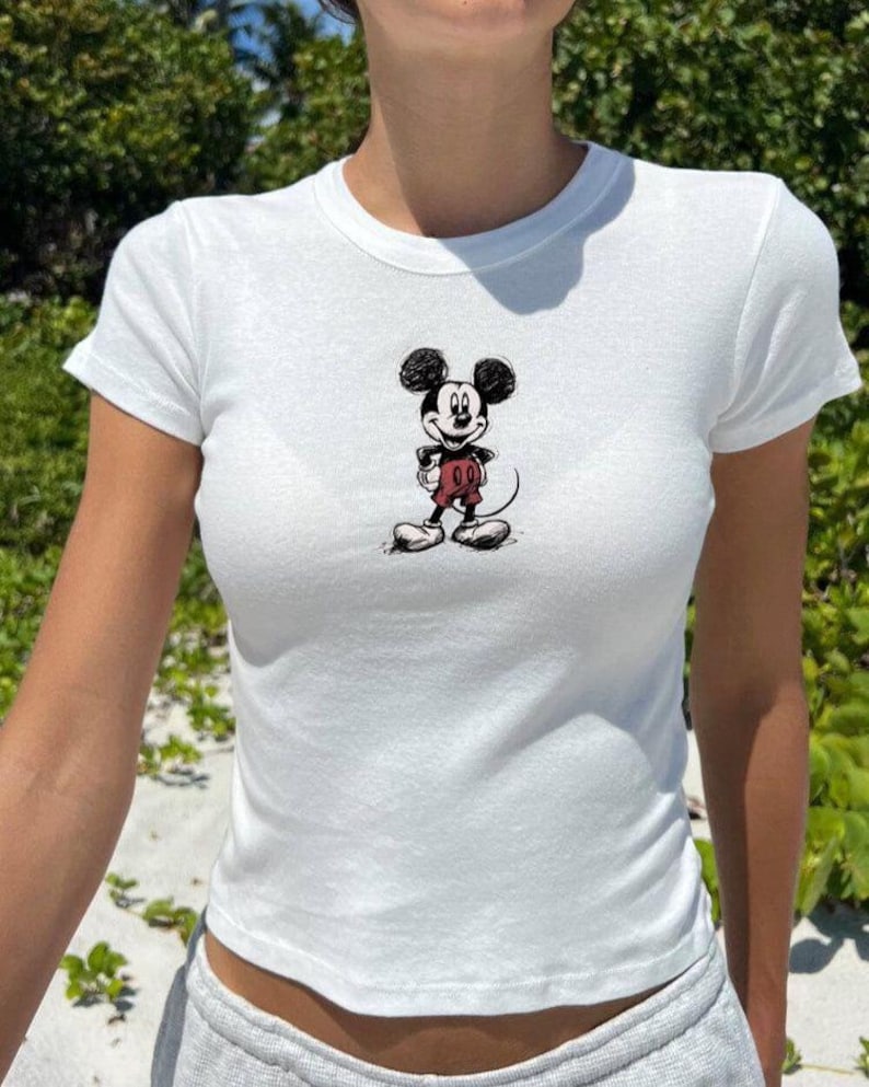 May include: White, short-sleeved t-shirt featuring a sketch-style Mickey Mouse graphic on the front. The cartoon character is depicted in black, red, and white. The shirt is made of a soft, lightweight material.