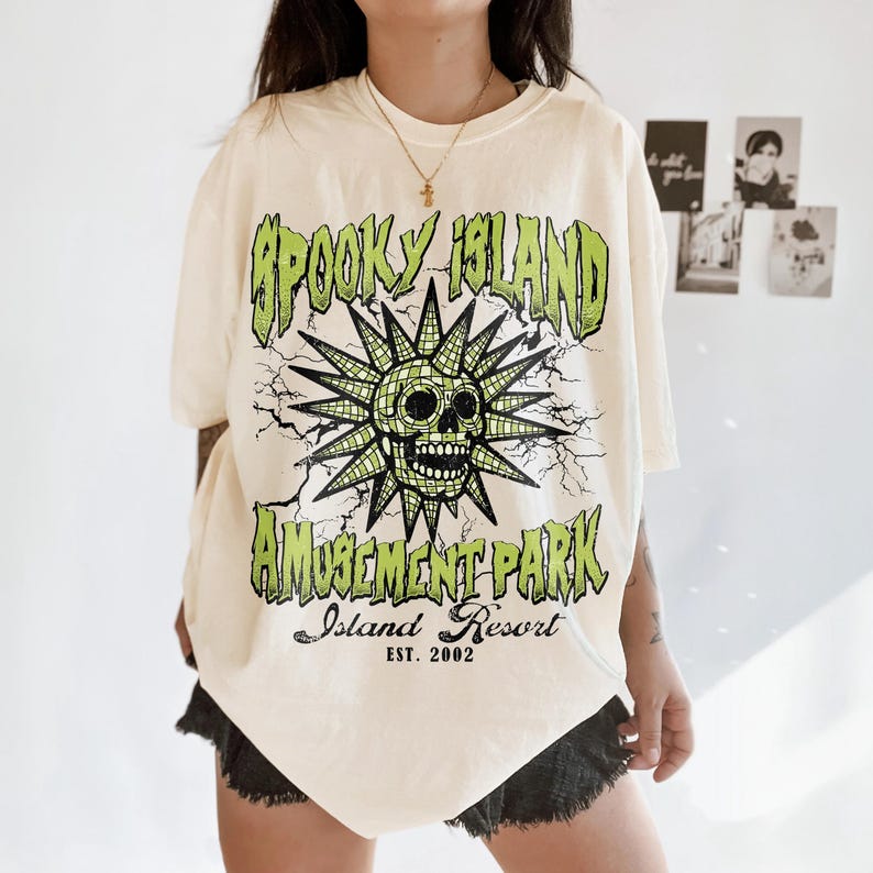 May include: Cream-colored t-shirt with a green and black graphic. The graphic features a skull with a sunburst design and the words "Spooky Island Amusement Park Island Resort EST. 2002".