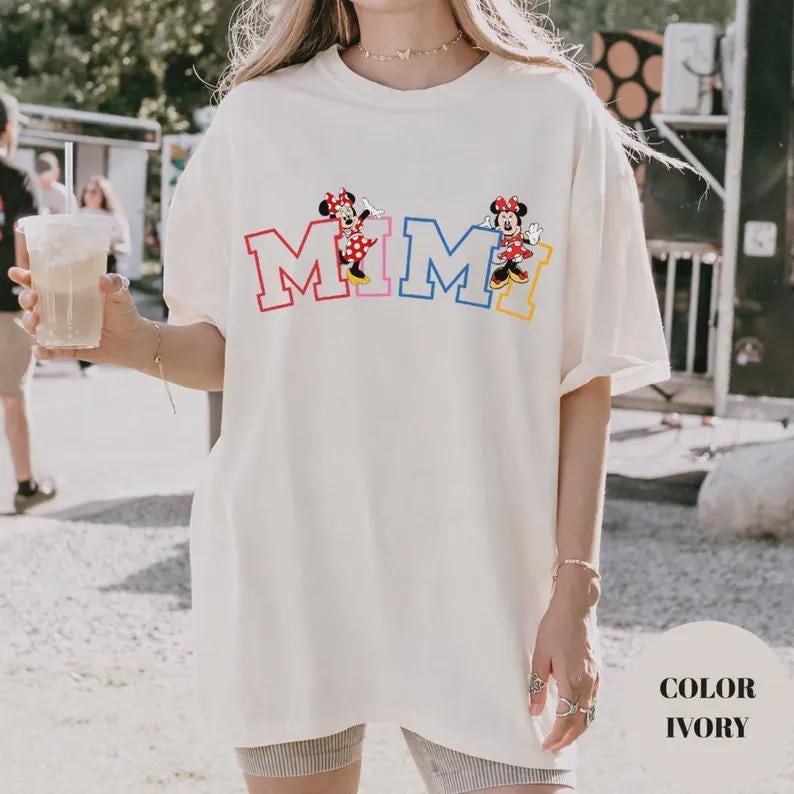 K&ouml;nnte beinhalten: Elfenbeinfarbenes T-Shirt mit dem Wort "MIMI" in bunten Blockbuchstaben und Minnie Mouse-Illustrationen. Das Shirt hat eine lockere Passform und kurze &Auml;rmel. Der Text "COLOR IVORY" befindet sich unten rechts.