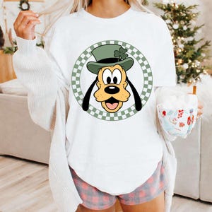 Peut inclure: Sweat-shirt blanc avec un dessin de Pluto portant un chapeau vert et un trèfle. Le dessin est encadré dans un cercle à carreaux verts et blancs. Fait partie d'une collection de vêtements.