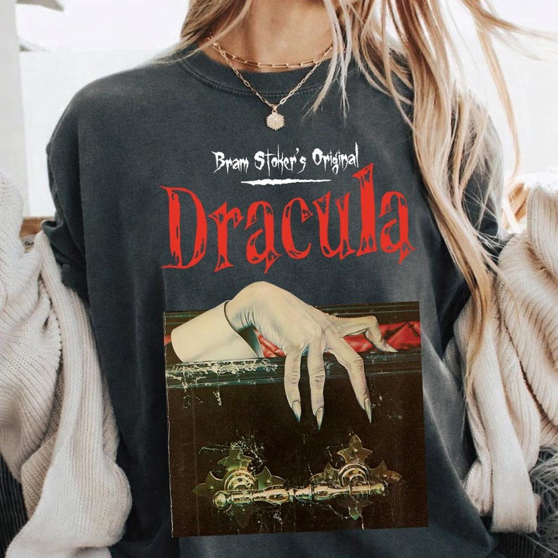 以下が含まれることがあります： 赤文字で「Bram Stoker's Original Dracula」と書かれたチャコールグレーのTシャツ。テキストの下には、鋭い爪を持つ青白い長い指の手が、金色の金具が付いた暗い木製の棺の縁に置かれているグラフィックが描かれています。