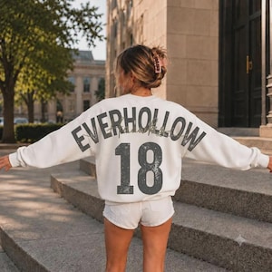 May include: A white long-sleeve crewneck sweatshirt featuring the text EVERHOLLOW in a large, distressed dark grey font arched across the upper back, with the number 18 printed in a matching large, distressed dark grey font below the text.