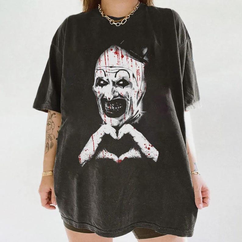 May include: Dark gray t-shirt featuring a graphic of a scary clown with blood splatter and a heart shape made with hands. The clown has white face paint, a black hat, and a sinister grin. The shirt is oversized.