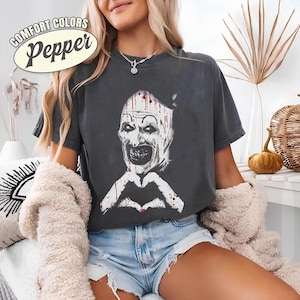 May include: Dark gray t-shirt featuring a white and red graphic of a scary clown face with a heart-shaped hand gesture. The text "COMFORT COLORS Pepper" is printed on the top left corner of the shirt.