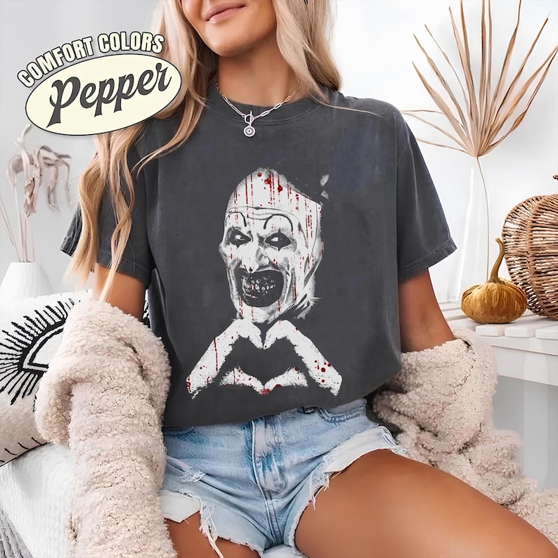 May include: Dark gray t-shirt featuring a white and red graphic of a scary clown face with a heart-shaped hand gesture. The text "COMFORT COLORS Pepper" is printed on the top left corner of the shirt.