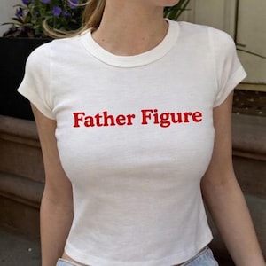 May include: White, short-sleeved t-shirt with the red text "Father Figure" across the chest. The shirt is a fitted style. The model is wearing blue jeans.