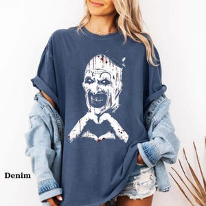 May include: A denim blue t-shirt featuring a white and black graphic of a scary clown face with a heart-shaped hand gesture. The word "Denim" is printed in the lower left corner of the image.