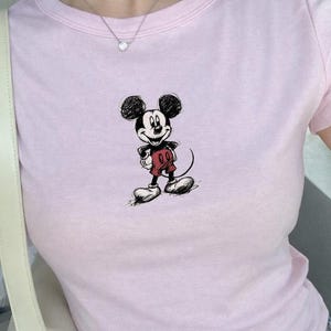 May include: A light pink t-shirt featuring a sketch of Mickey Mouse. The cartoon character is depicted in black, red, and white. The shirt has a crew neck and short sleeves. The overall style is casual and suitable for everyday wear.