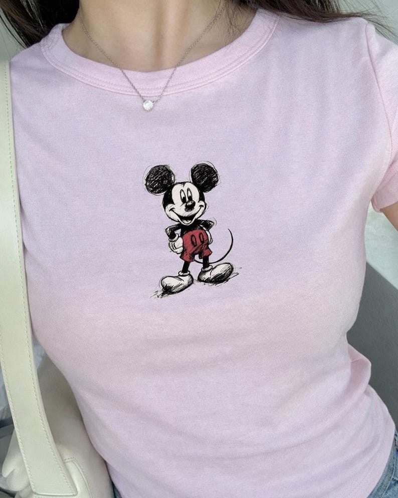 May include: A light pink t-shirt featuring a sketch of Mickey Mouse. The cartoon character is depicted in black, red, and white. The shirt has a crew neck and short sleeves. The overall style is casual and suitable for everyday wear.