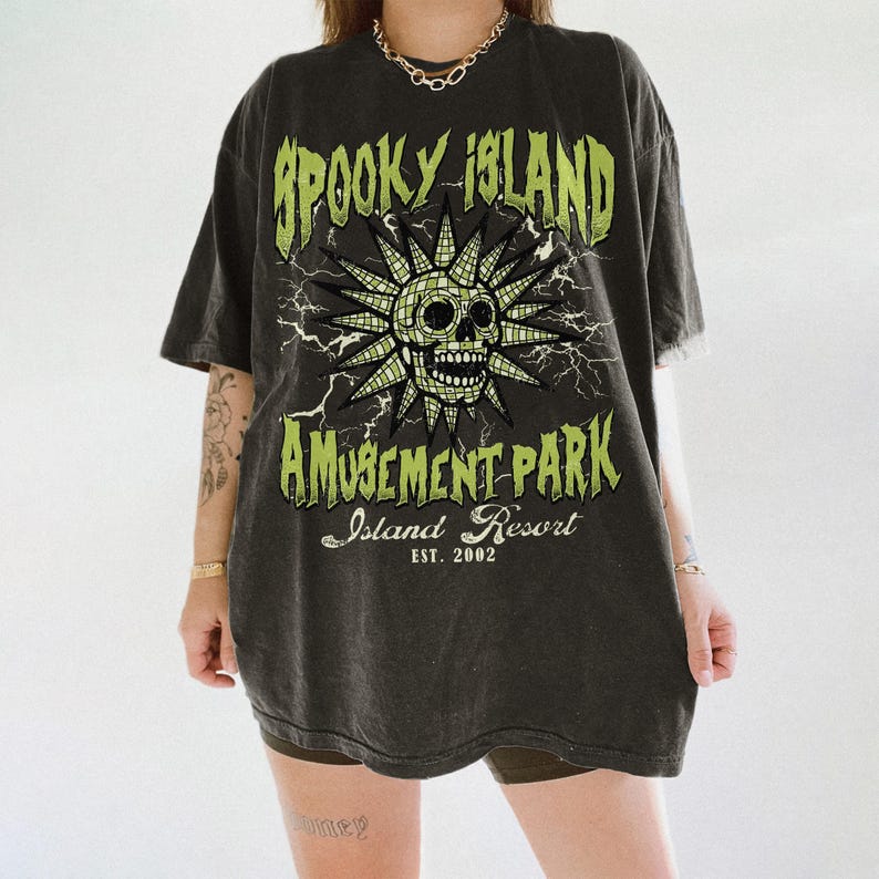 May include: Dark gray t-shirt with a distressed graphic design. The design features a skull with a sunburst, lightning bolts, and the words "Spooky Island Amusement Park Island Resort EST. 2002" in green.