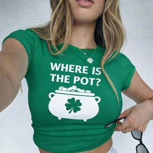 May include: A green t-shirt with the text "WHERE IS THE POT?" in white. Below the text is a white graphic of a pot of gold with a four-leaf clover. The shirt has short sleeves and a crew neck.