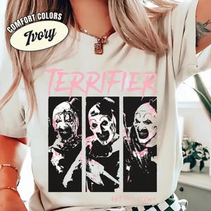 May include: An ivory short-sleeve t-shirt featuring a graphic print with the text "TERRIFIER" in pink above three black and white panels showing Art the Clown. The text "ART THE CLOWN" appears in small pink letters at the bottom of the graphic.