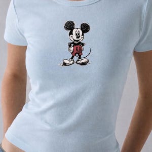 May include: Light blue t-shirt featuring a sketch of Mickey Mouse. The cartoon character is depicted in black and white with red shorts and white gloves and shoes. The shirt is a crew neck style.