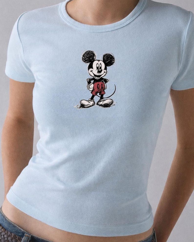 May include: Light blue t-shirt featuring a sketch of Mickey Mouse. The cartoon character is depicted in black and white with red shorts and white gloves and shoes. The shirt is a crew neck style.