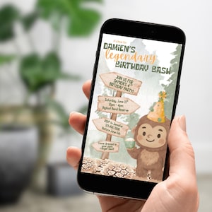 May include: A smartphone displays a birthday party invitation. The invitation features a cartoon Bigfoot holding a cupcake, wearing a party hat, and surrounded by wooden signs with event details. The text reads "Damien's Legendary Birthday Bash."