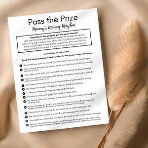 May include: A white printed game card titled "Pass the Prize" with the subtitle "Mommy's Morning Mayhem." The card includes instructions for a game and a story about a busy morning. The card is on a beige surface.