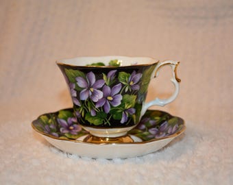 May include: A white ceramic mug with a black handle and the text "But First, Coffee" in a bold, black font. The mug is set against a blurred background, suggesting a cozy setting.