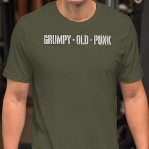 Puede incluir: Una camiseta de manga corta color verde oliva con el texto blanco "GRUMPY • OLD • PUNK" impreso en el pecho. La camiseta tiene cuello redondo y un corte estándar, confeccionada con una tela de color sólido y un diseño sencillo.
