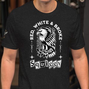 May include: Dark gray t-shirt featuring a black and white eagle design with the text "RED, WHITE & BROKE" and "Squiggy". The shirt is short-sleeved and has a crew neck. The person is standing in a gym.