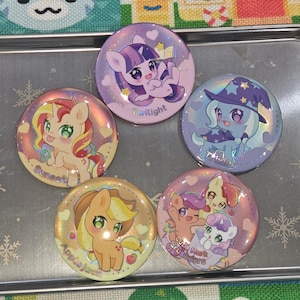 May include: A collection of five round, colorful pinback buttons featuring cartoon pony characters. Each button has a different character with names like "Twilight", "Sunset", and "Applejack". The buttons have a glossy finish and are set on a silver tray.