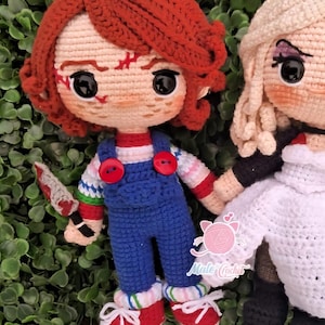Könnte beinhalten: Gehäkelte Puppe einer Figur mit roten Haaren, blauer Latzhose und einem Spielzeugmesser. Die Puppe hat ein genähtes Gesicht und trägt rot-weiß gestreifte Ärmel und rote Schuhe. Daneben eine weitere gehäkelte Puppe mit blonden Haaren und einem weißen Kleid.