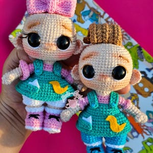 May include: Two handmade crochet dolls. One doll has a pink bow, the other has a brown hat. Both dolls have large black eyes, eyelashes, and are wearing teal overalls with yellow duck appliques, pink shirts, and pink and white shoes. The dolls are holding hands.