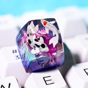 May include: A custom keycap featuring a koi fish design. The keycap is a translucent resin with a purple, blue, and white color scheme. The koi fish are white with black markings and red accents. The keycap is on a white keyboard.
