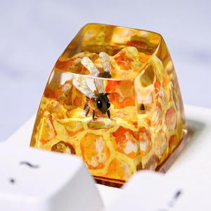 Może przedstawiać: Niestandardowa nakładka na klawiaturę z pszczołą w przezroczystej żywicy. Nakładka ma wzór plastra miodu w odcieniach żółci i pomarańczy. Pszczoła jest czarno-żółta. Nakładka znajduje się na białej klawiaturze.