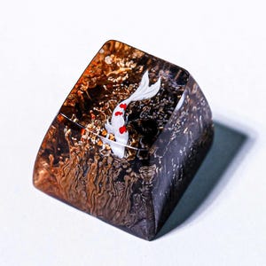 May include: A custom artisan keycap featuring a white koi fish with red spots swimming in a clear resin block. The resin has a brown and gold textured appearance, creating a miniature aquatic scene. The keycap is angled and sits on a white surface.