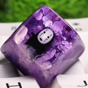 Può includere: Un copritasto personalizzato con un design in resina viola raffigurante il personaggio Senza Volto di Spirited Away. Il copritasto di forma quadrata è posizionato su una tastiera bianca. La resina ha un aspetto vorticoso e strutturato.