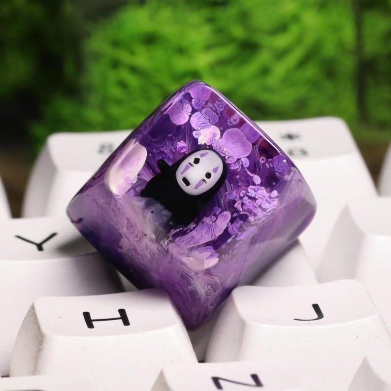 May include: A custom keycap featuring a purple resin design with a No-Face character from Spirited Away. The keycap is square-shaped and sits atop a white keyboard. The resin has a swirling, textured appearance.