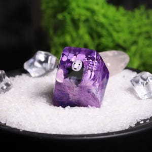 May include: A purple resin keycap featuring a character from Spirited Away. The keycap has a No-Face design, with a black body and white mask. The keycap is surrounded by white crystals and sits on a bed of white sand.