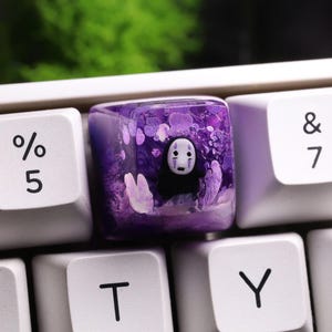 May include: A custom keyboard keycap featuring a purple resin design with a character from Spirited Away. The keycap is square and sits among other white keyboard keys. The character is black and white with a white mask.