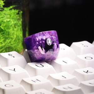May include: A custom purple keycap featuring a character from Spirited Away. The keycap has a translucent design with a white and black character in the center. The keycap is resting on a white keyboard.