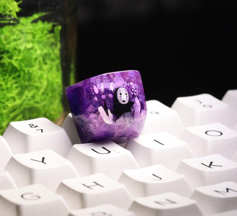 May include: A custom purple keycap featuring a character from Spirited Away. The keycap has a translucent design with a white and black character in the center. The keycap is resting on a white keyboard.