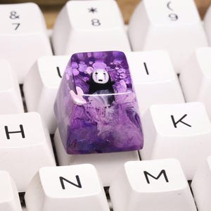 May include: A custom keycap featuring a No-Face character from Spirited Away. The keycap is a vibrant purple with a swirling, marbled effect. The No-Face figure is black with a white mask. The keycap is surrounded by white keyboard keys.