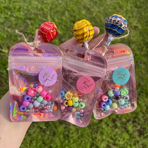 May include: Three iridescent pink resealable pouches containing assorted colourful plastic beads, including flower-shaped and round beads. Each pouch is tied with twine to a round lollipop in red, yellow, or blue, and features a thank you sticker.