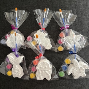 May include: Six clear plastic bags containing white plaster shapes including a seashell, seahorse, starfish, mermaid tail, jellyfish, and turtle. Each bag includes a small paintbrush and a strip of small paint pots with various colours.