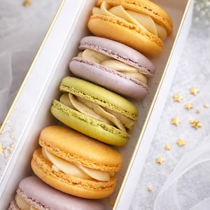 May include: A box of colorful macarons, including yellow, lavender, and green, filled with creamy white filling. The macarons are arranged in a white box with gold trim, set against a soft, light background.