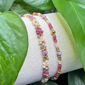 May include: Two beaded bracelets. One features small, pink flower-shaped beads and the other has pink and gold beads. The bracelets are displayed on a white surface, surrounded by green leaves.